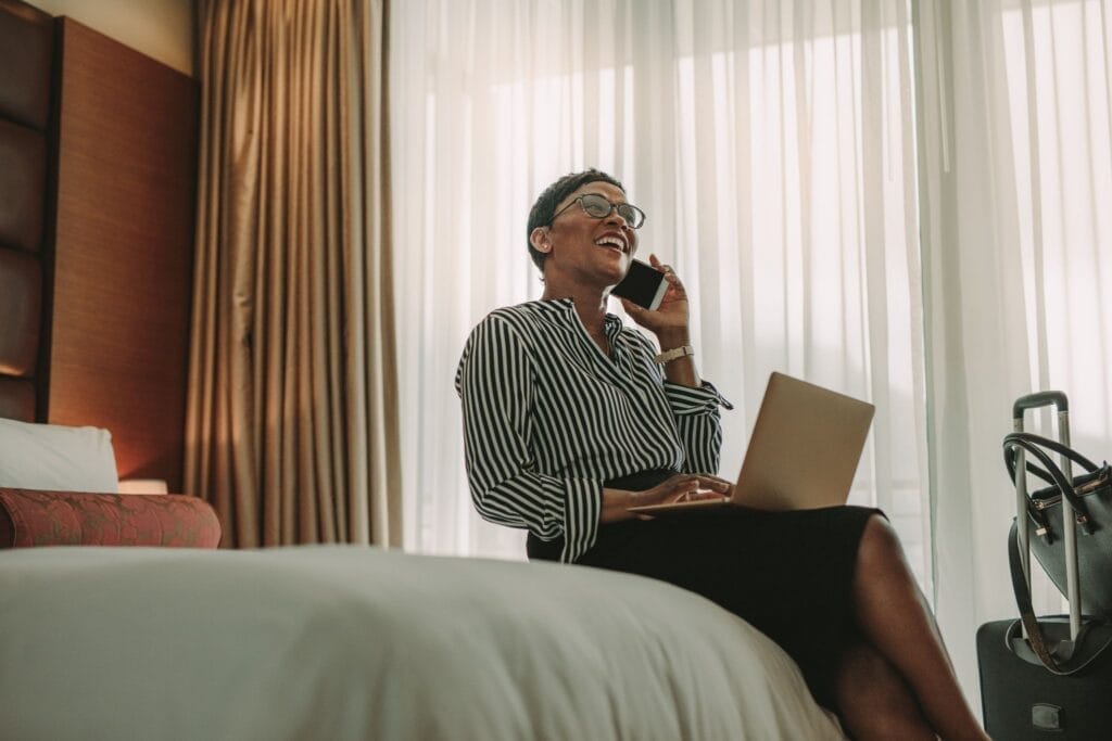 Hotel guest smiling and talking on the phone while sitting on her hotel room bed.