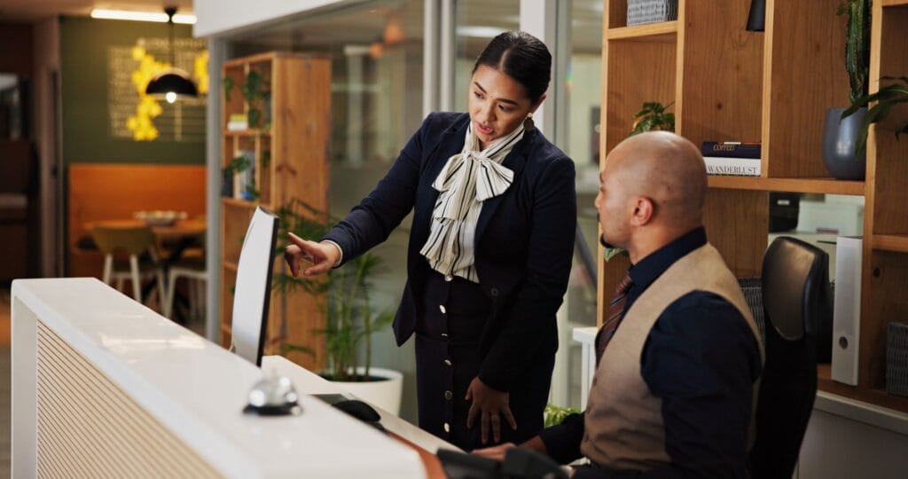 Two hotel employees talking to each other at the front desk. One is pointing to something on the computer.